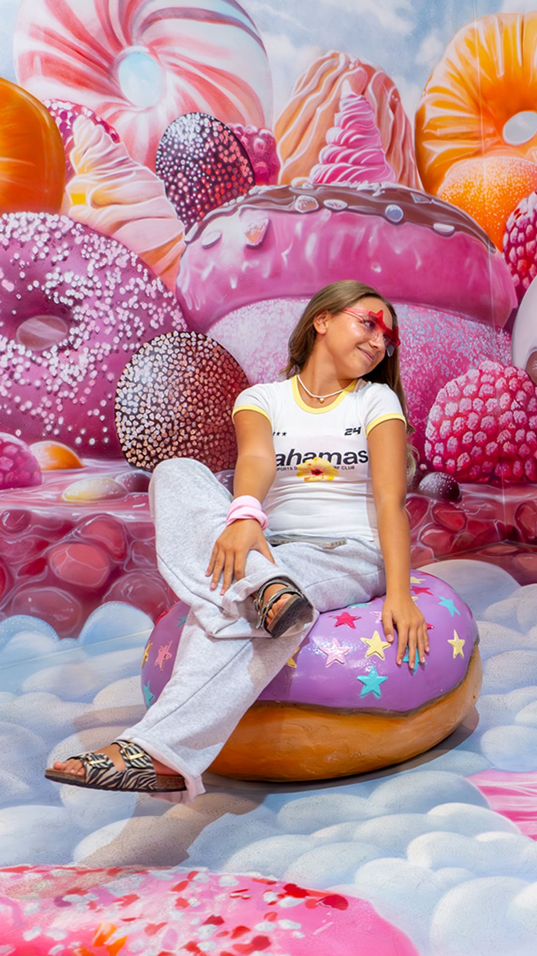Two young visitors sitting on giant donut seats in front of a towering wall of oversized candy, strawberries and cakes at Sweet Museum Barcelona