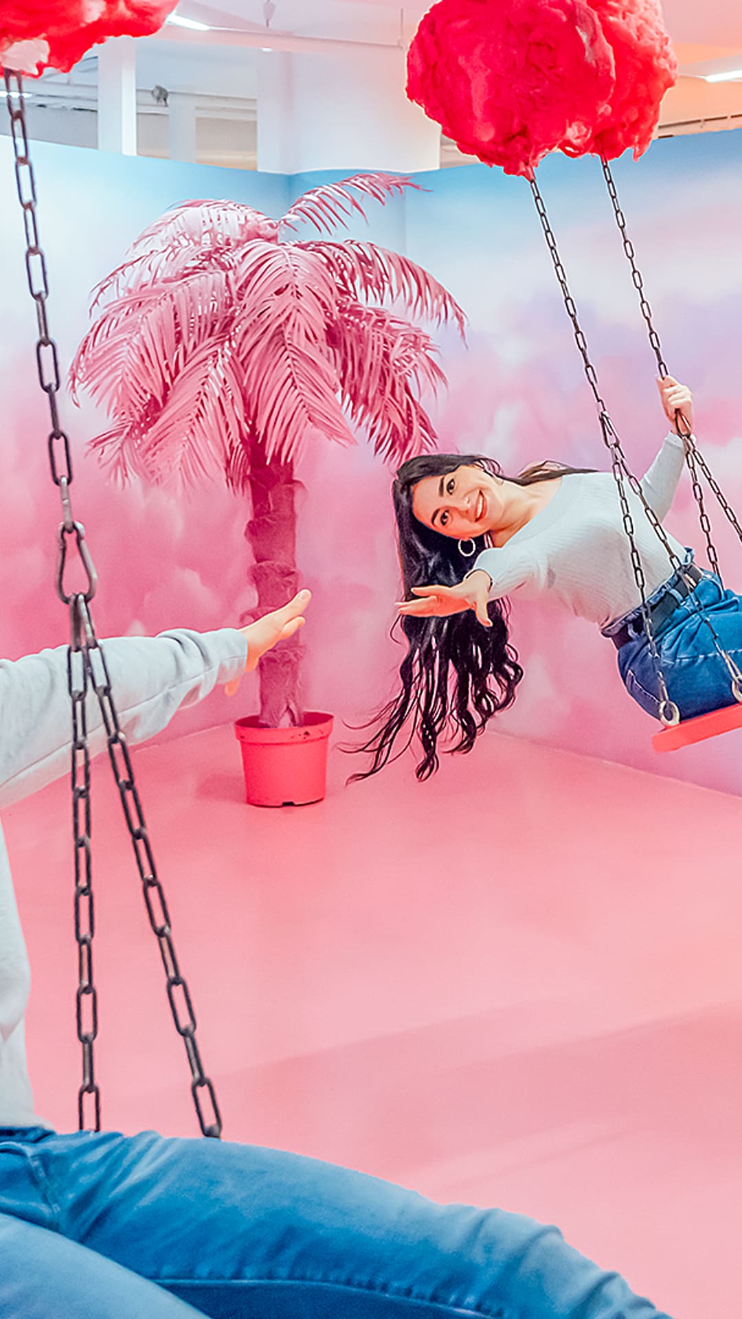 A boy and a girl swinging from fluffy pink cotton-candy clouds in a pastel pink room with a pink palm tree and giant donuts at Sweet Museum Barcelona