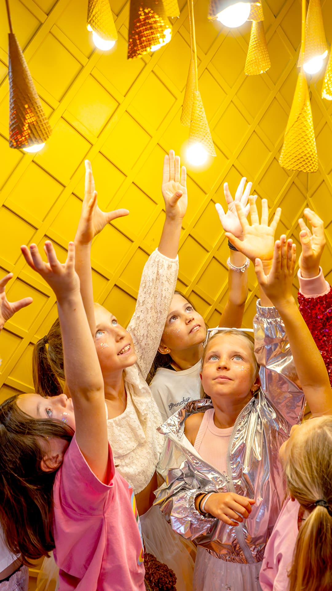 A group of children reaching up toward glowing ice-cream-cone lights on a bright yellow ceiling at Sweet Museum Barcelona